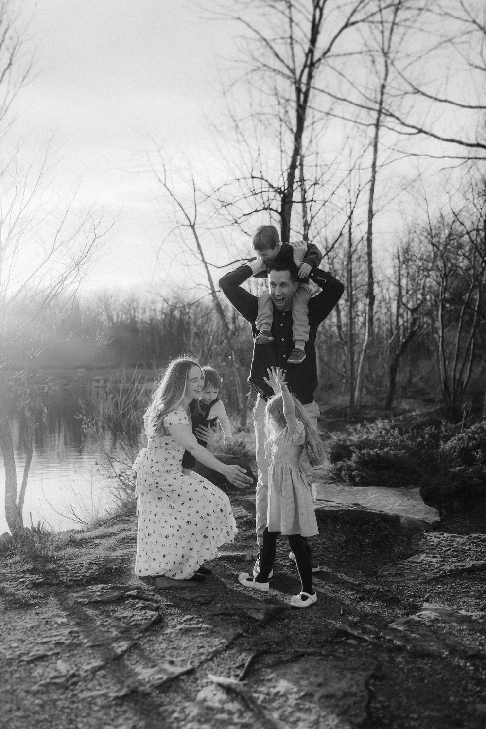 A family enjoys a playful moment together by a serene lake on an overcast day in black and white.