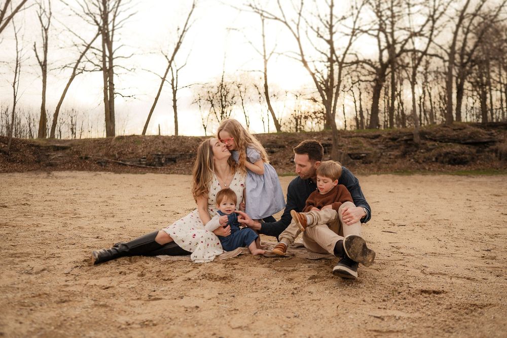 A family sits together on sandy ground at sunset with bare trees in the background.
