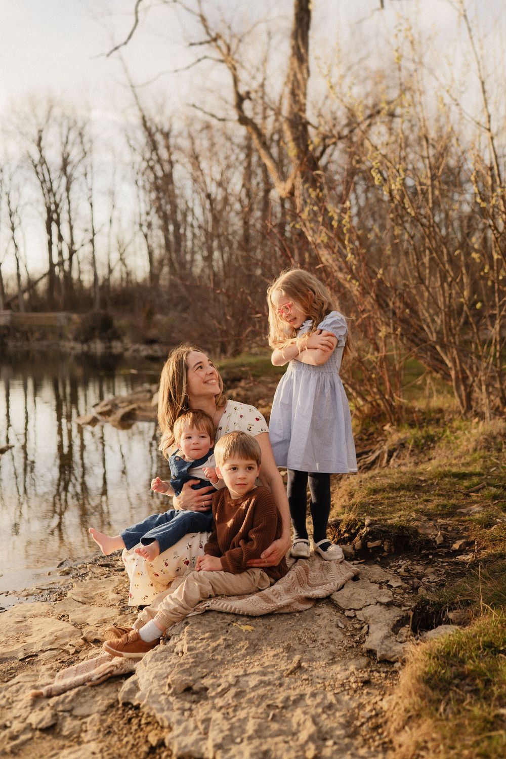 A family sits together on rocks by a tranquil pond surrounded by winter trees in warm evening light.