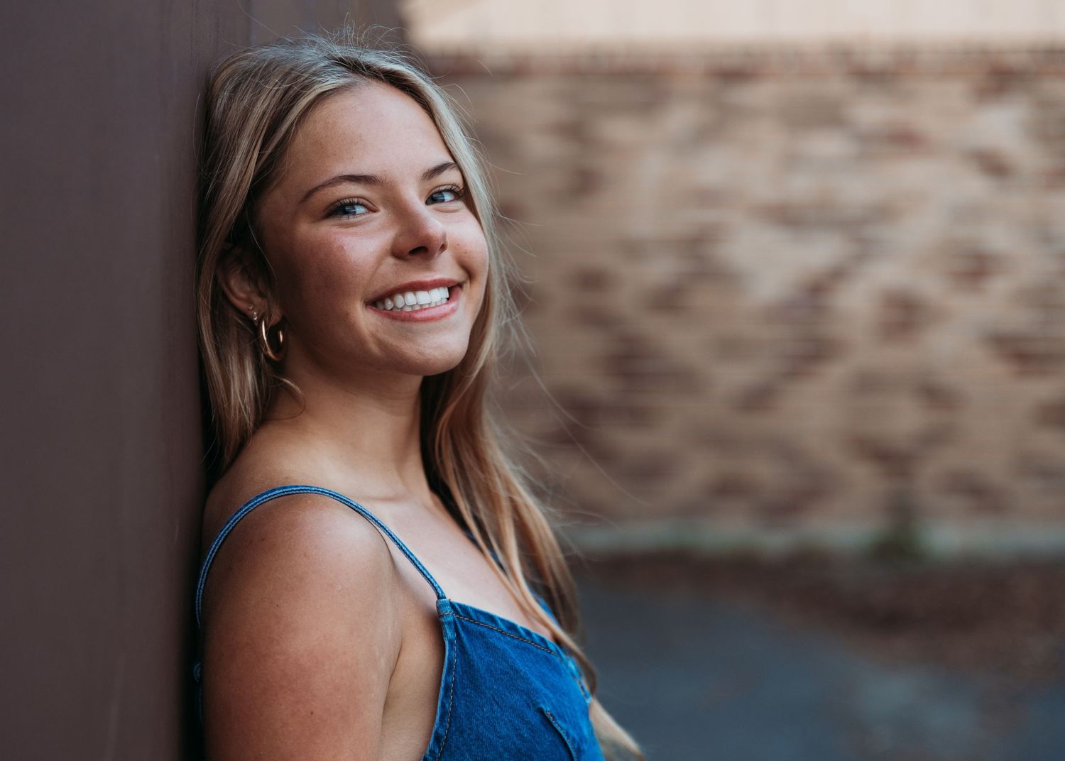 A person in a blue sleeveless top smiling brightly against a rustic brick wall background.