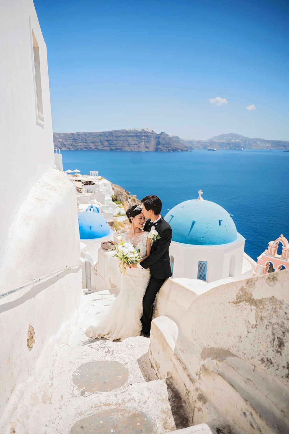 Wedding couple posing against iconic white buildings and blue domes of Santorini, Greece.