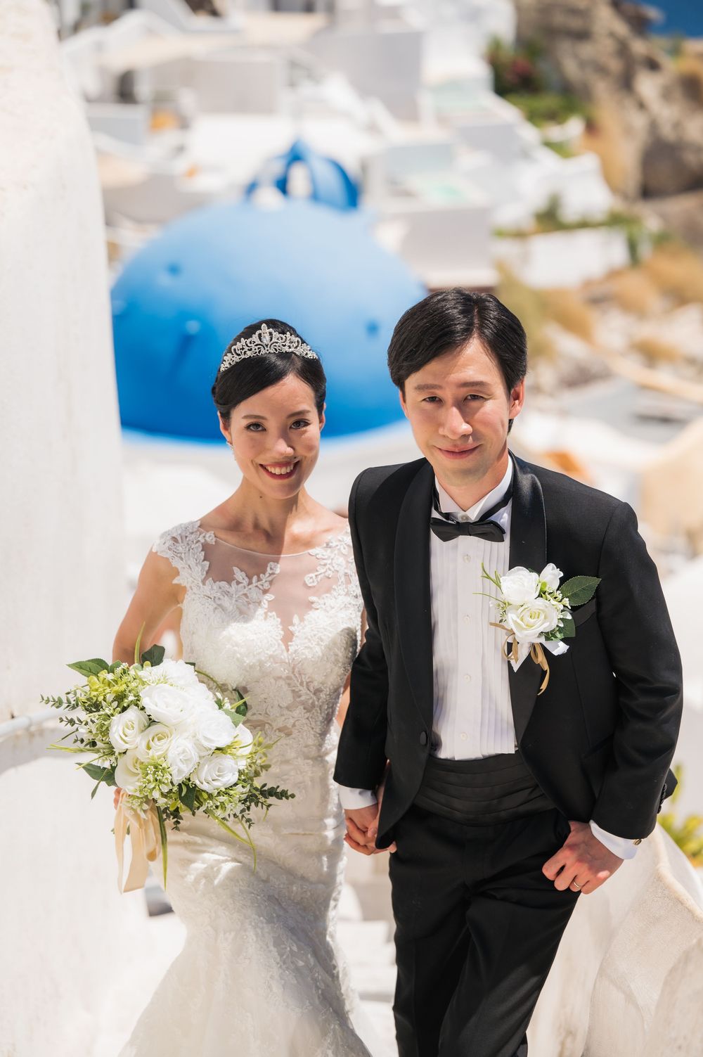 Wedding couple in formal attire posing on white steps with Santorini's blue domes in background.