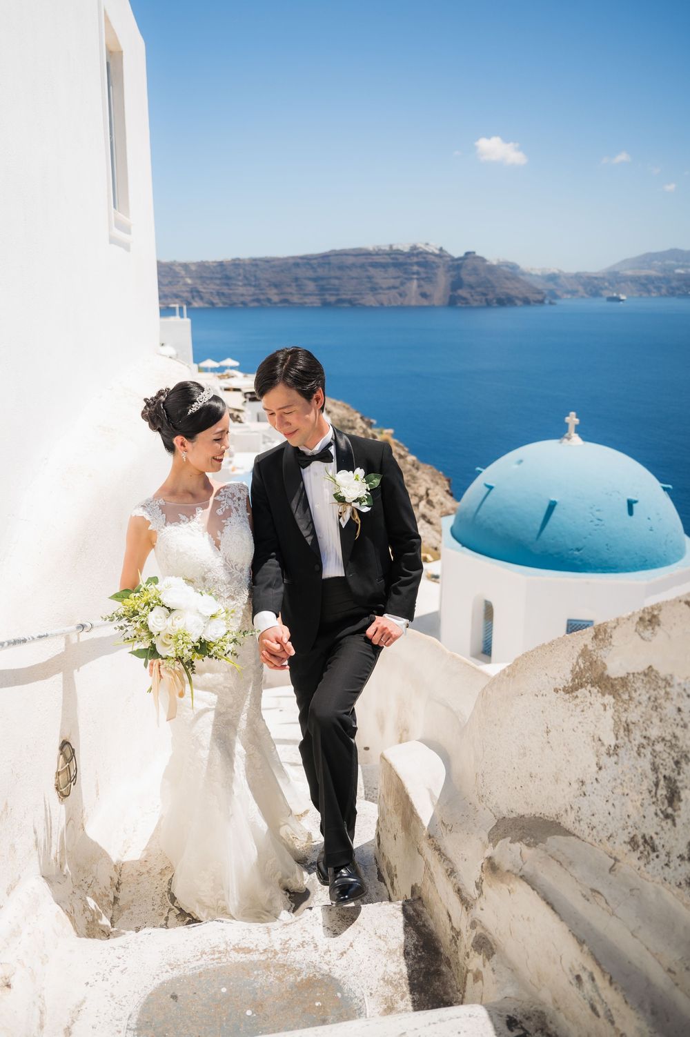 Wedding couple sharing intimate moment on white steps with iconic Santorini blue dome and sea view.