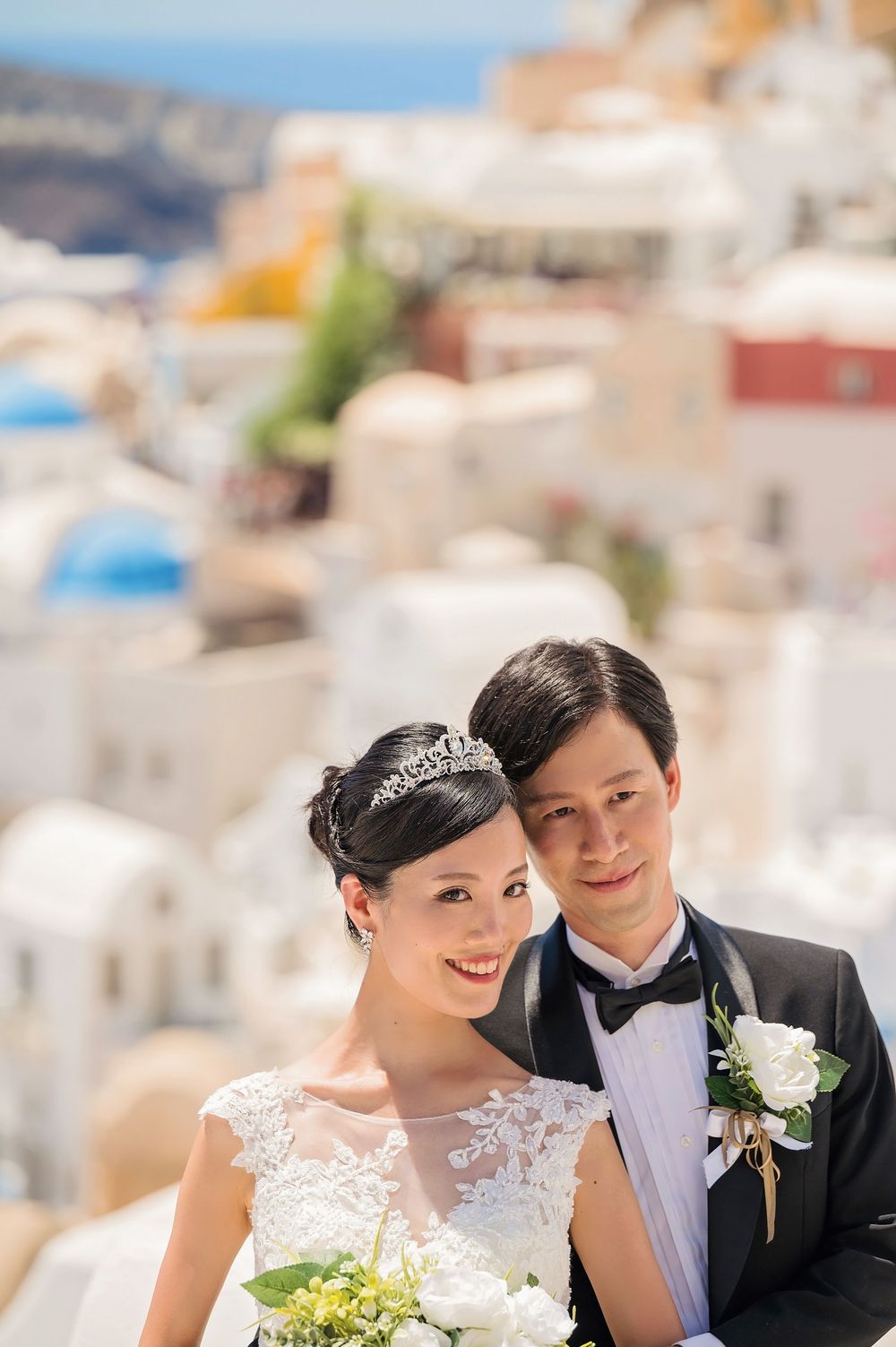 A sunlit wedding portrait captured in Santorini with white cycladic architecture in the background.