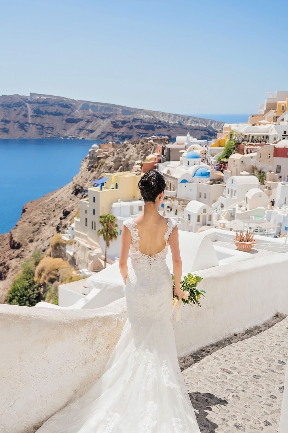 A bride in a fitted lace gown overlooking the stunning Santorini caldera and white village landscape.