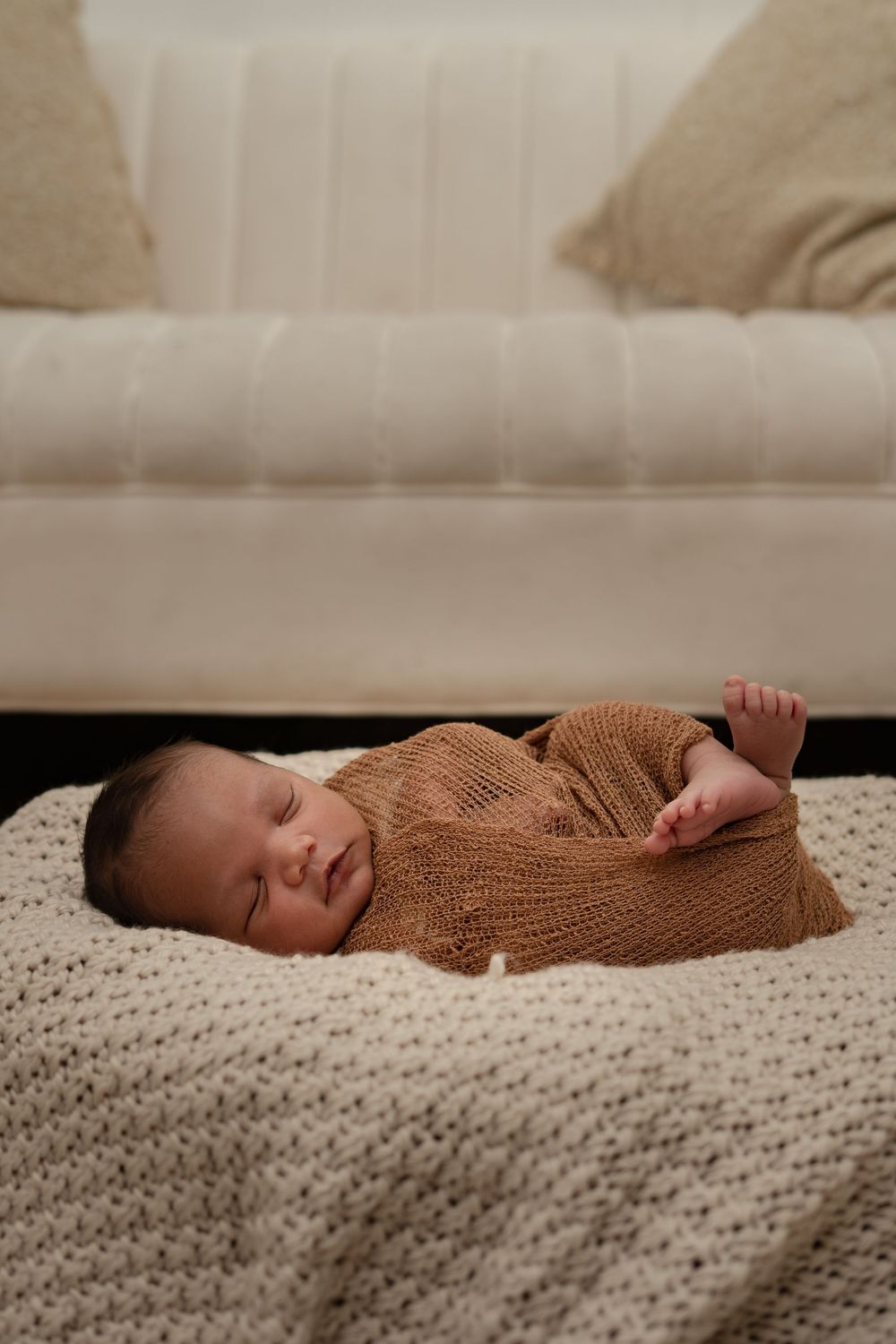 Sleeping newborn wearing brown knit outfit on cream textured blanket.