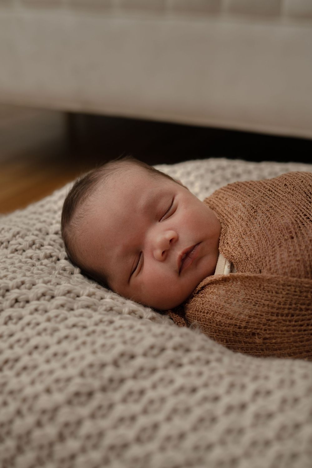 Newborn baby peacefully sleeping on textured blanket wrapped in soft brown swaddle.