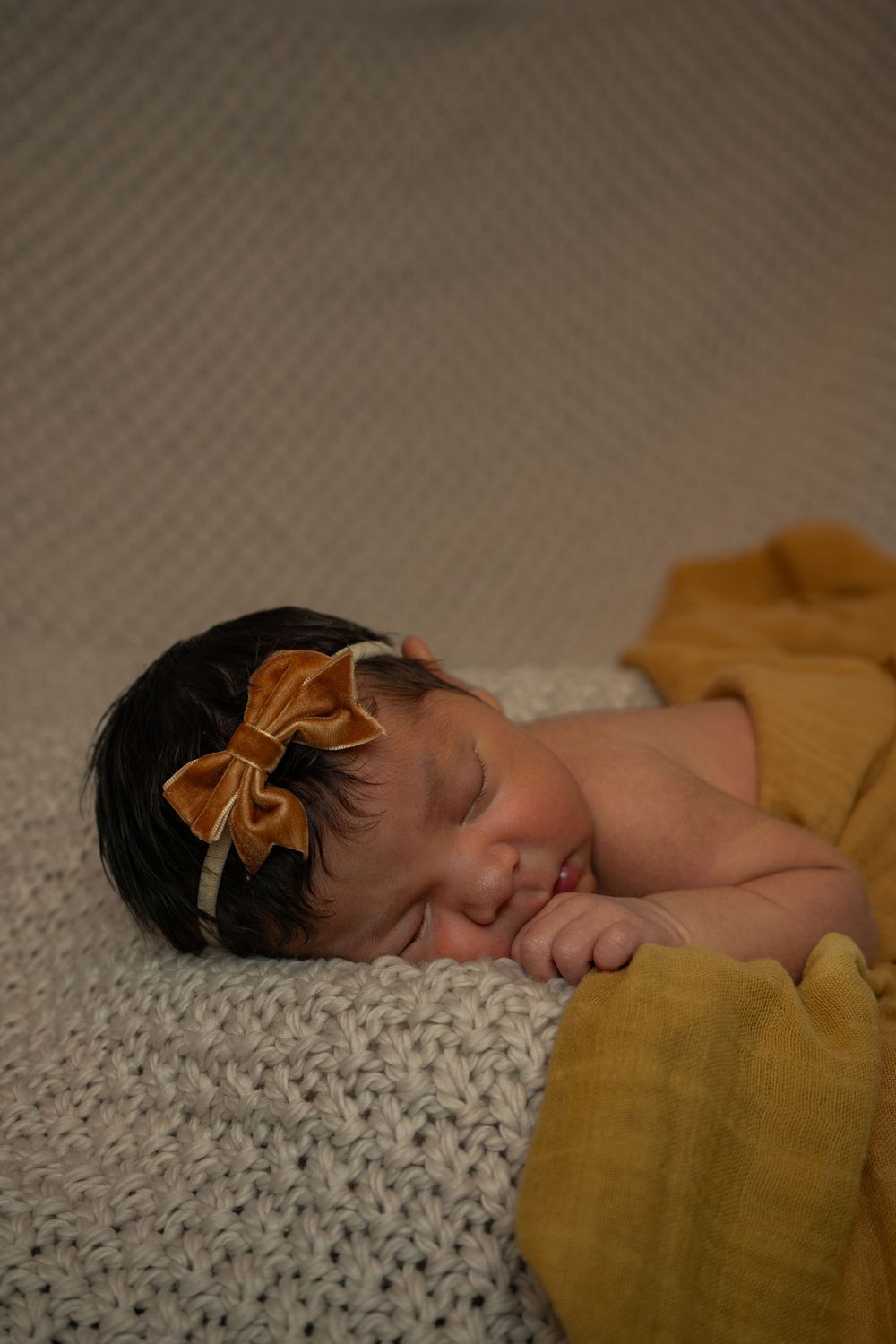 Sleeping newborn on textured blanket with mustard yellow fabric and bow accessory.