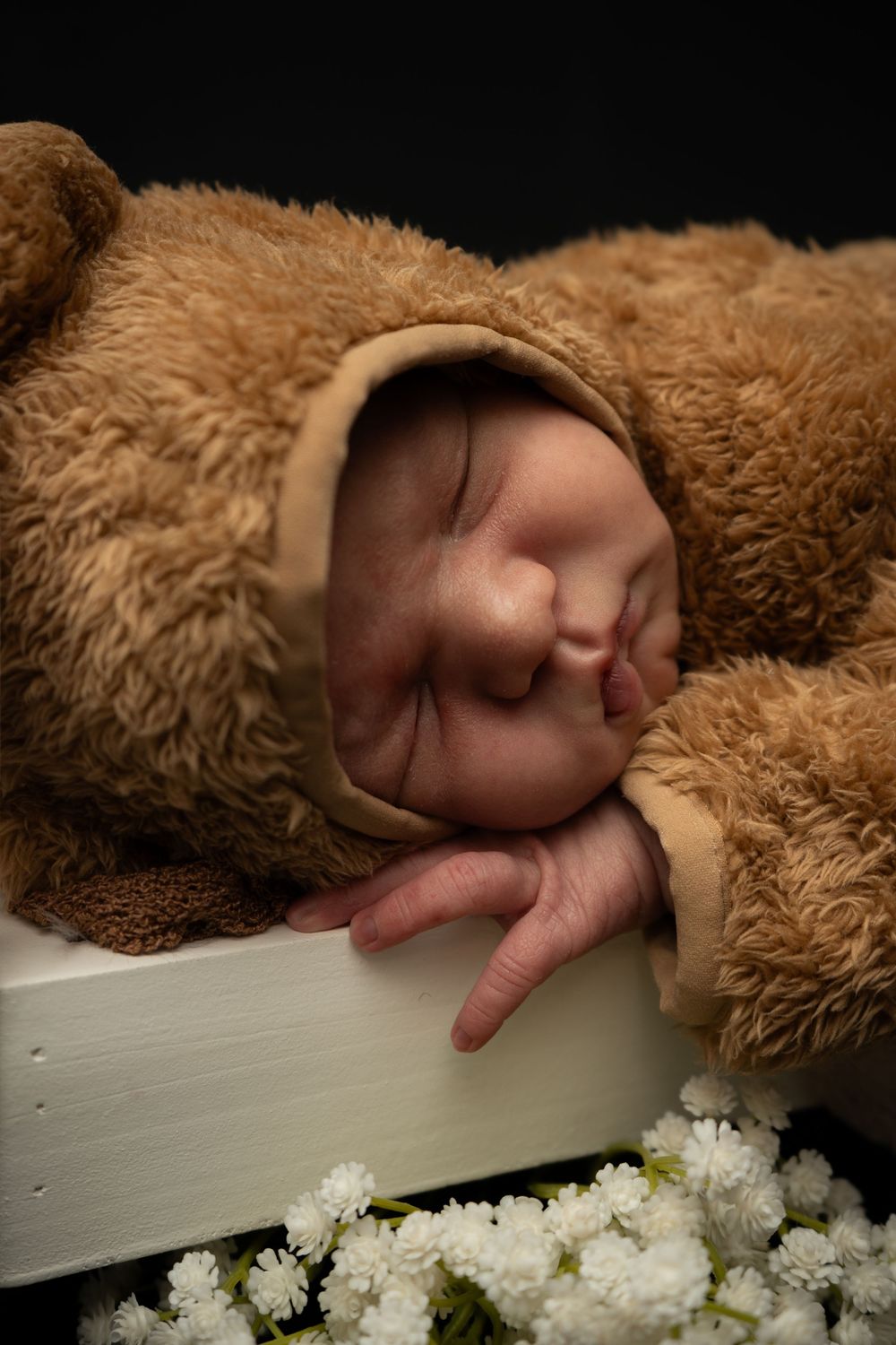 A peaceful baby nestled in a fuzzy brown teddy bear outfit sleeps beside delicate white flowers.