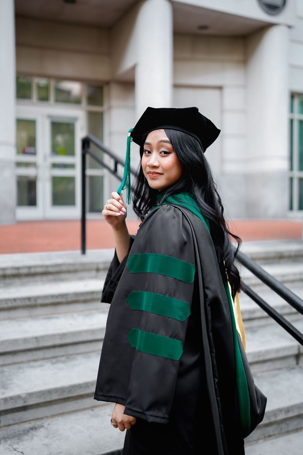 Young woman in black and green doctoral graduation gown and cap poses on university steps, smiling over shoulder, in Baltimore, MD