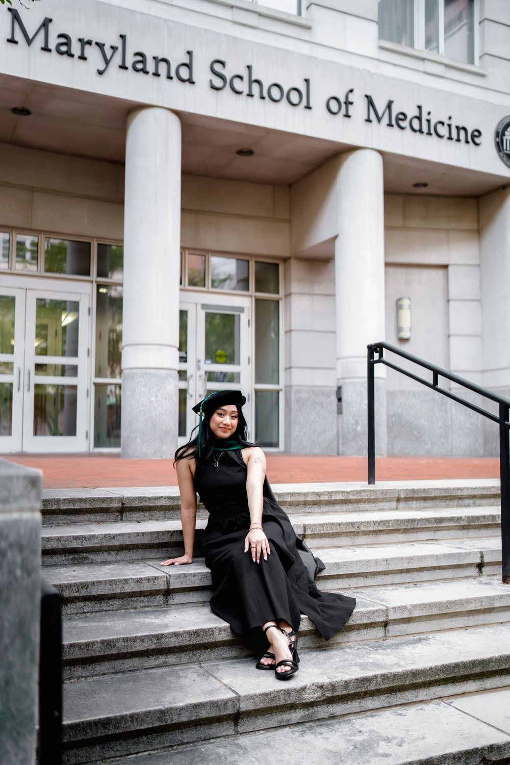 Graduate in black gown and cap sits on steps outside University of Maryland School of Medicine building, in Baltimore, MD