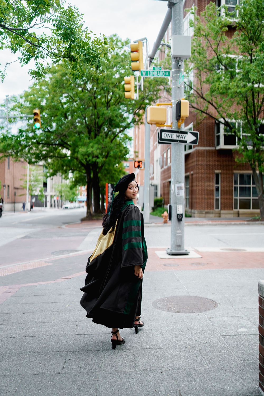 A graduate in cap and gown poses on a city sidewalk near street signs and traffic lights, surrounded by green trees, in Baltimore, MD