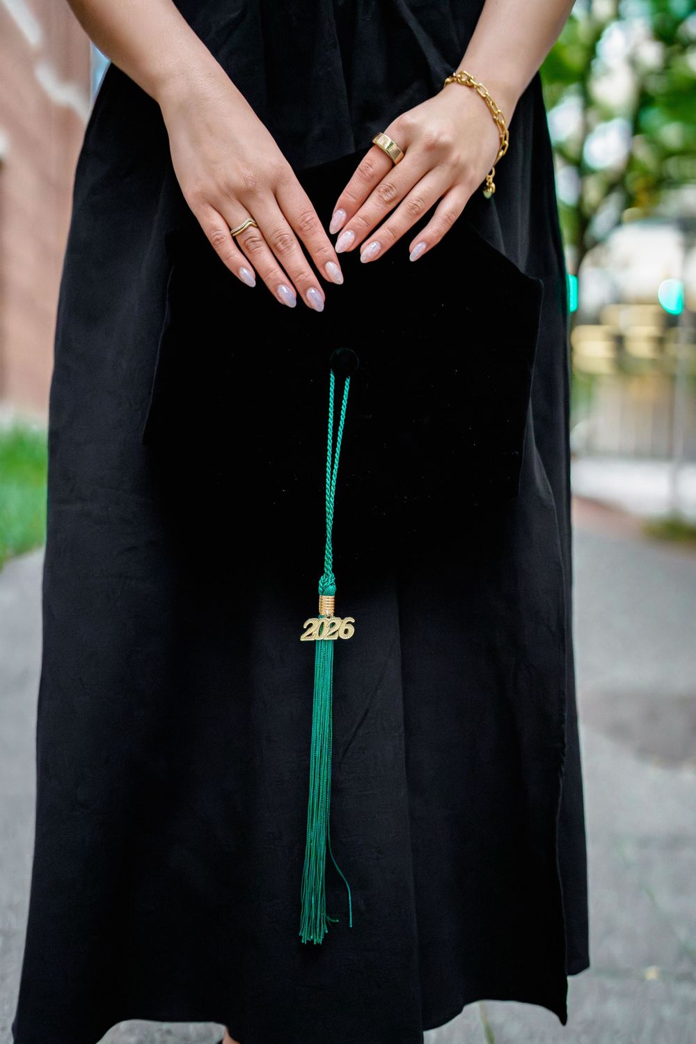 Graduate holding black cap with green 2026 tassel, wearing rings and gold bracelet in black graduation gown, University of Maryland, MD