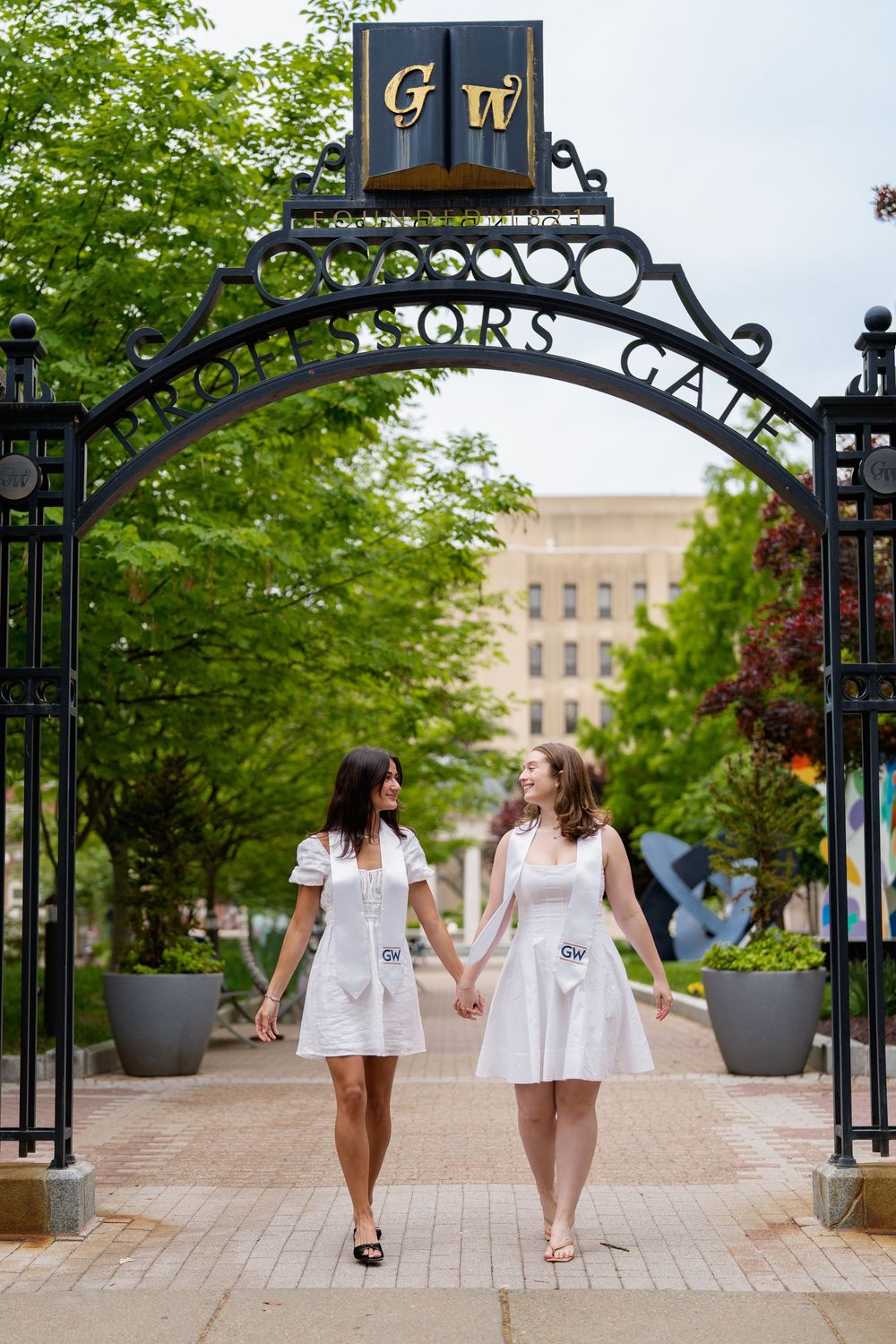 Two GW graduates in white dresses holding hands under the Scholars Gate arch, during graduation session in Washington DC