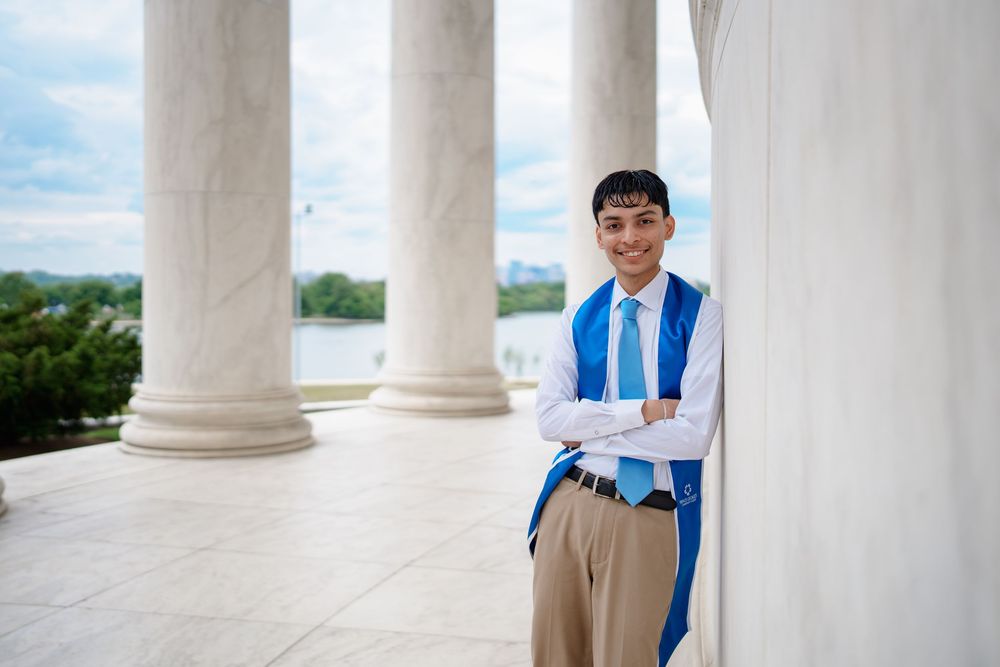 Young man in blue graduation sash poses confidently at Jefferson Memorial columns with Potomac River backdrop.