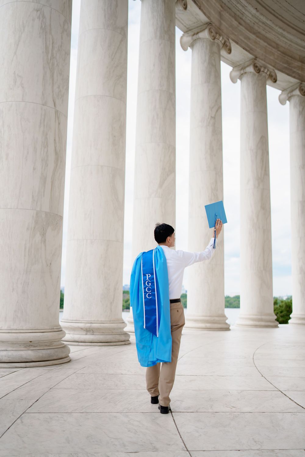 Graduate in blue regalia raises diploma among white marble columns at the Jefferson Memorial in Washington DC.
