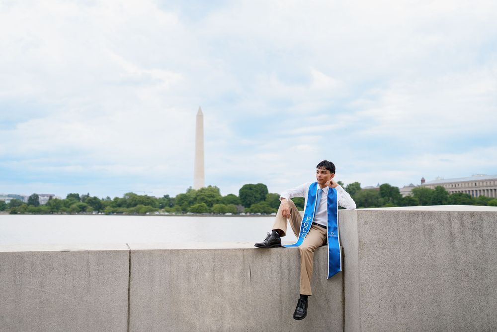 Graduate in cap and gown sits on concrete wall near Washington Monument and Tidal Basin in Washington DC.