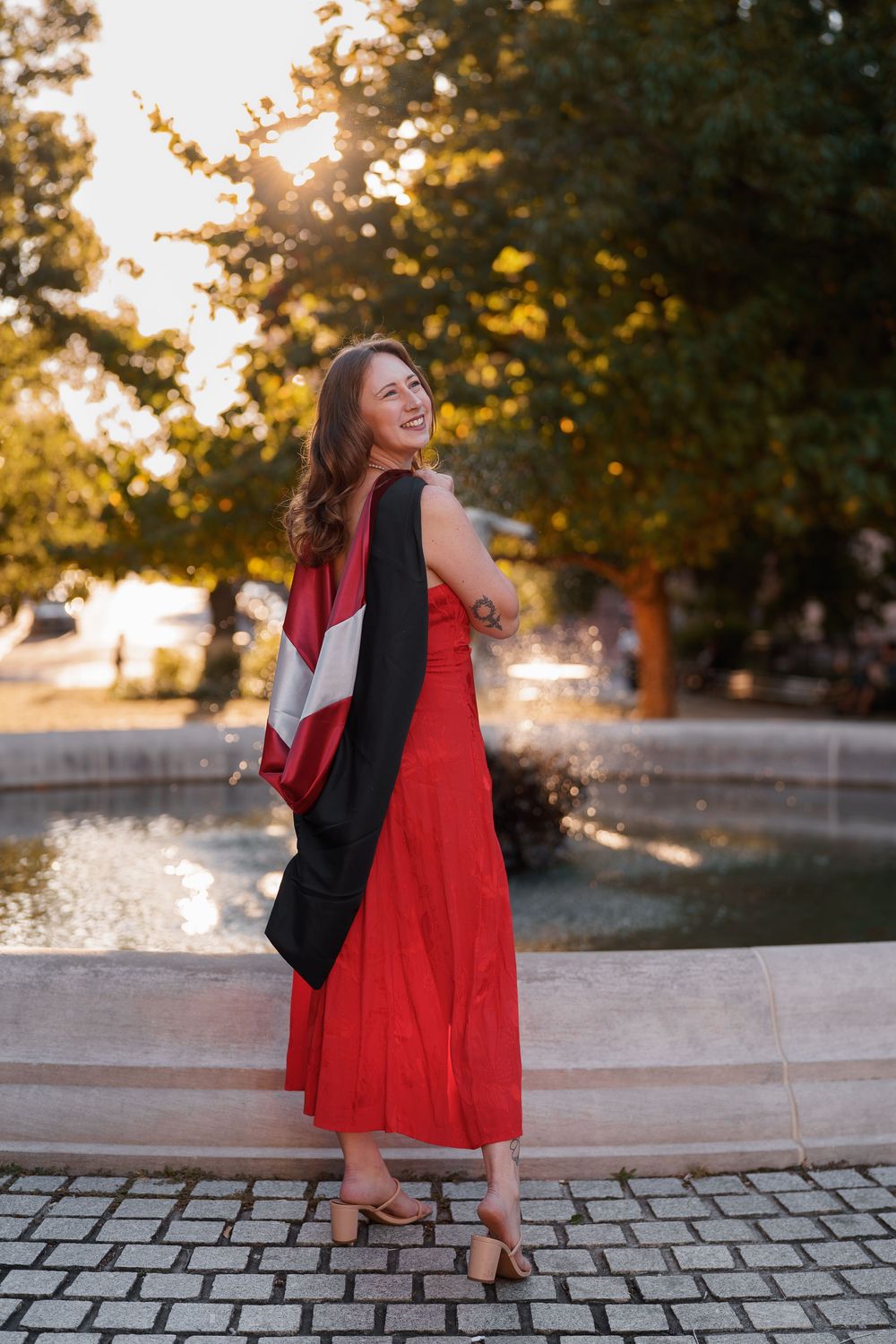 Woman in red dress twirls joyfully at sunset on stone pavement near riverfront with trees in background, in Baltimore, MD