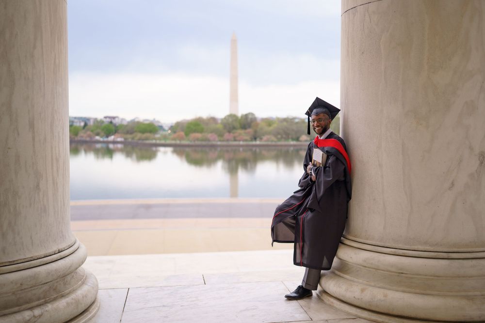 Graduate in cap and gown sits thoughtfully between white columns during a graduation photo session.