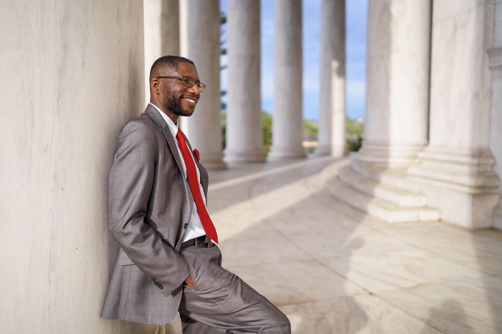 Professional Black businessman in gray suit and red tie smiling confidently while leaning against marble columns, in DC