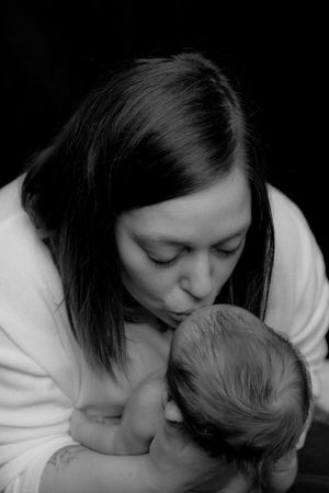A tender black and white portrait shows a parent gently kissing their baby's head while wearing a white sweater.