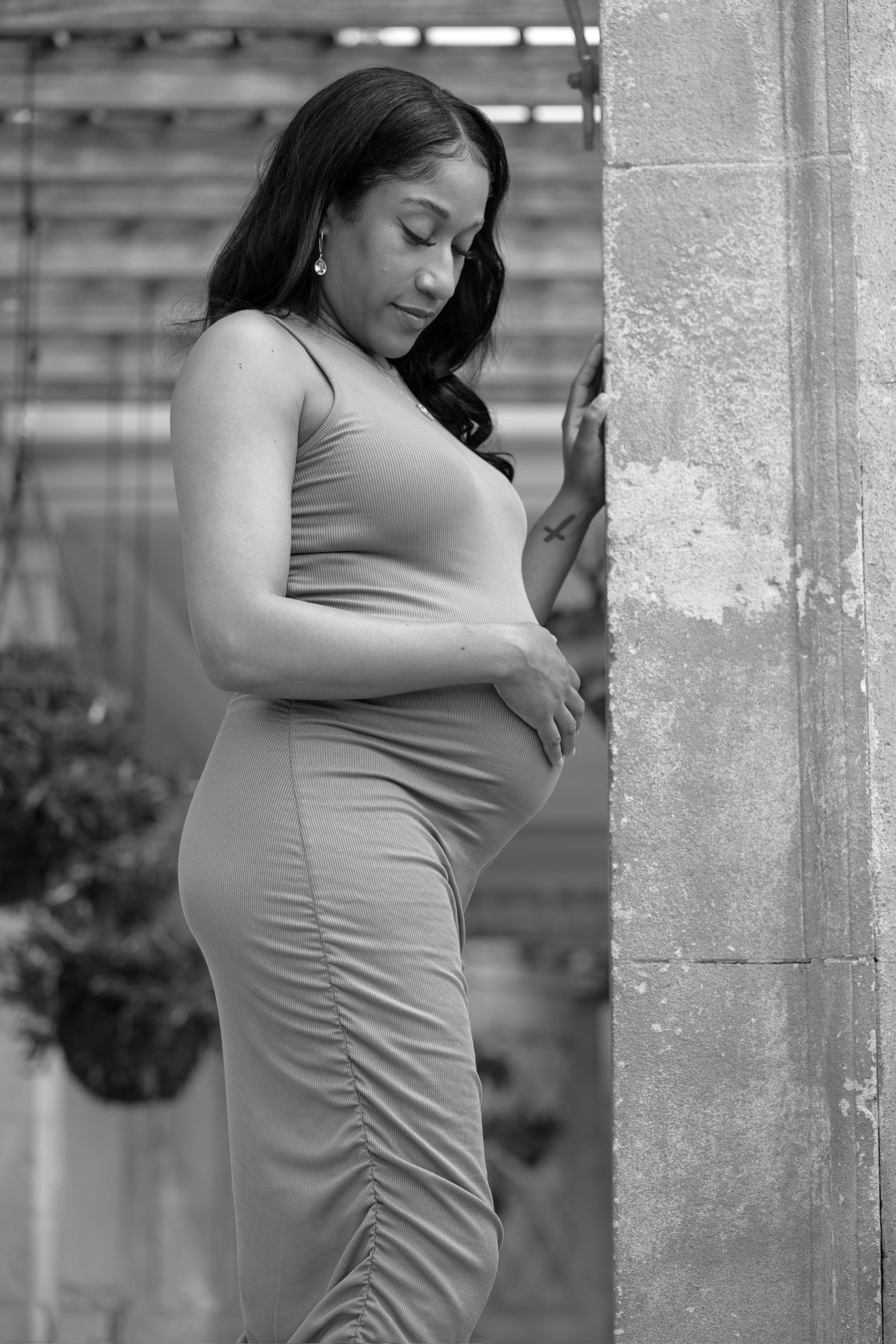 Black and white maternity portrait of pregnant woman in form-fitting dress leaning against stone wall.
