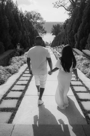 A couple in white outfits walking hand in hand down a tree-lined garden path on a sunny day.