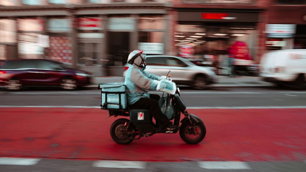 Delivery rider speeds past on motorcycle against blurred urban background.