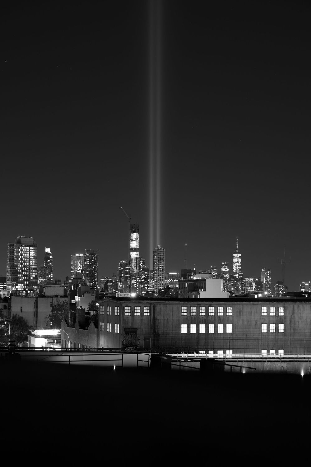 Twin beams of light illuminate the New York City skyline at night with silhouetted buildings in the foreground.