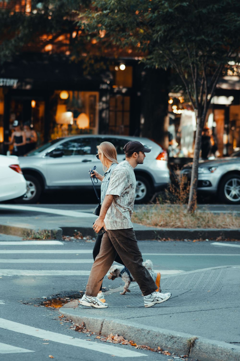 People crossing an urban street at dusk with parked cars and illuminated storefronts.