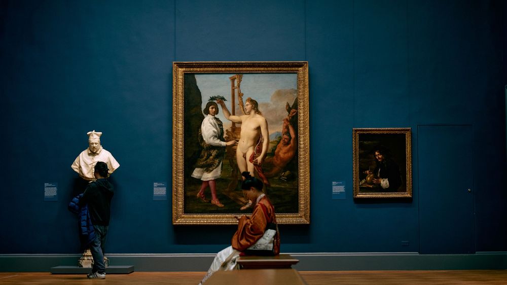 Museum visitors admire classical artwork in an elegant blue-walled gallery.