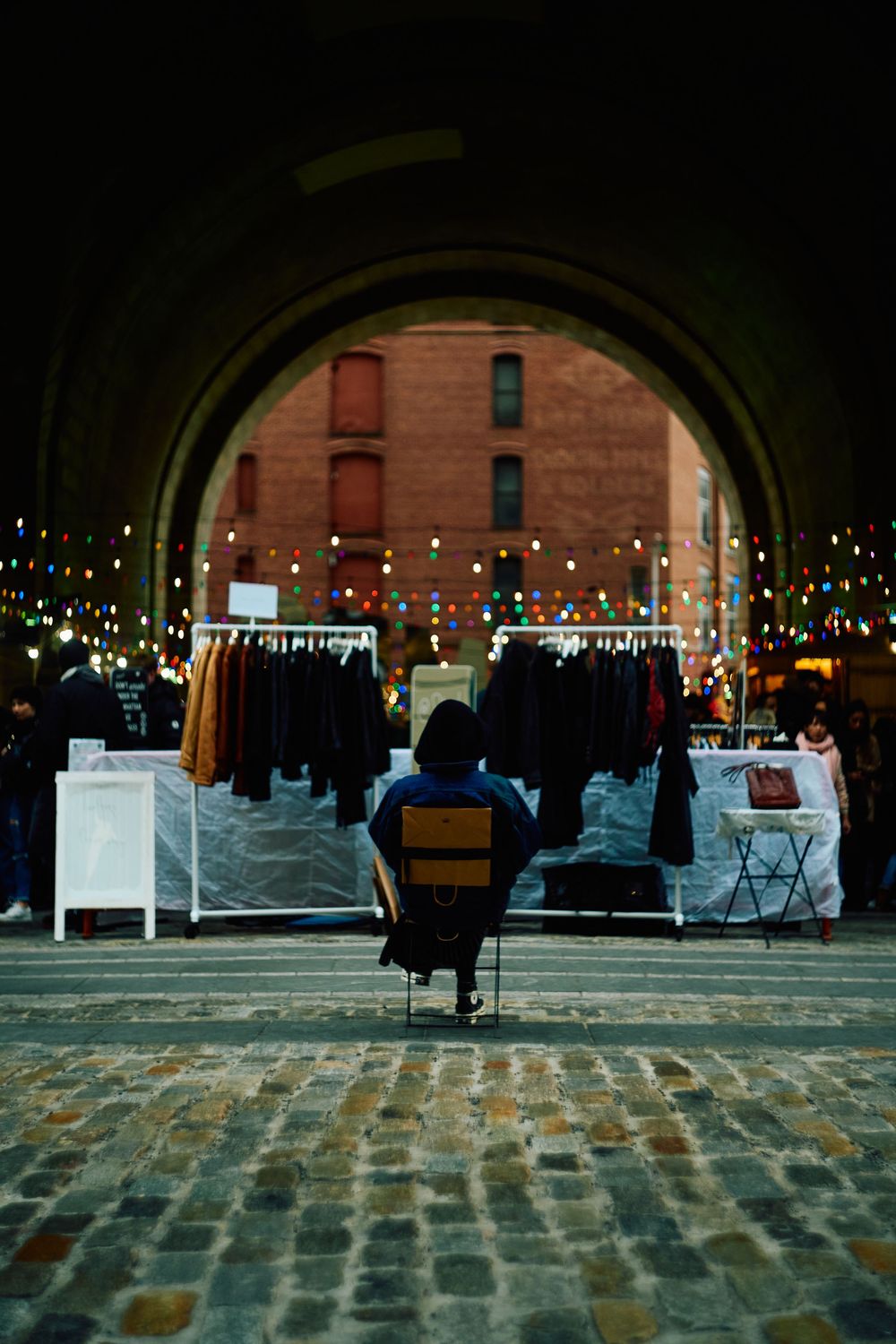 Vendor sits in archway surrounded by clothing displays at night market.