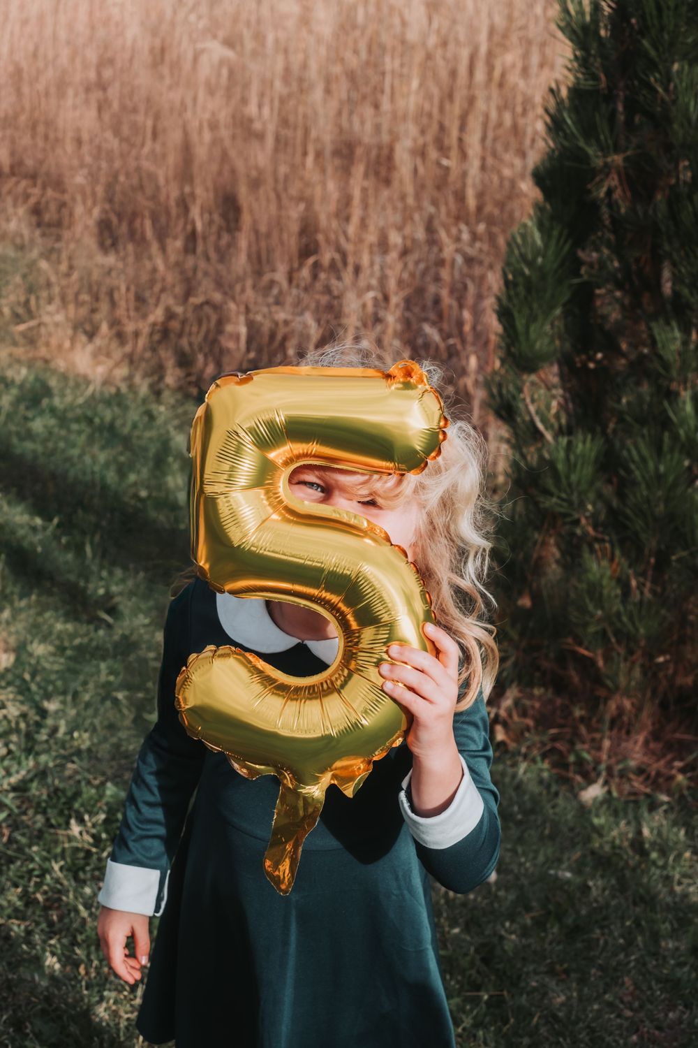 A young girl gives a silly face as she peeks through part of a number five balloon.
