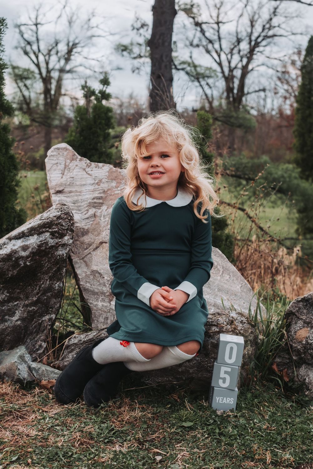 A young girl with wild blonde hair sits on a rock with blocks acknowledging her age next to her.