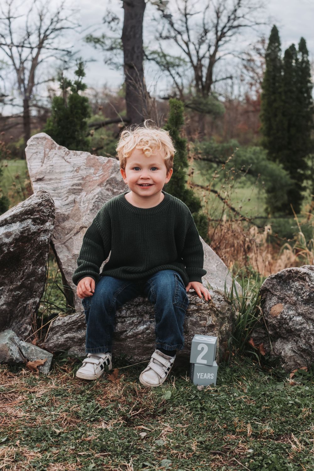A young boy sits on a rock with a block stating his age next to him.