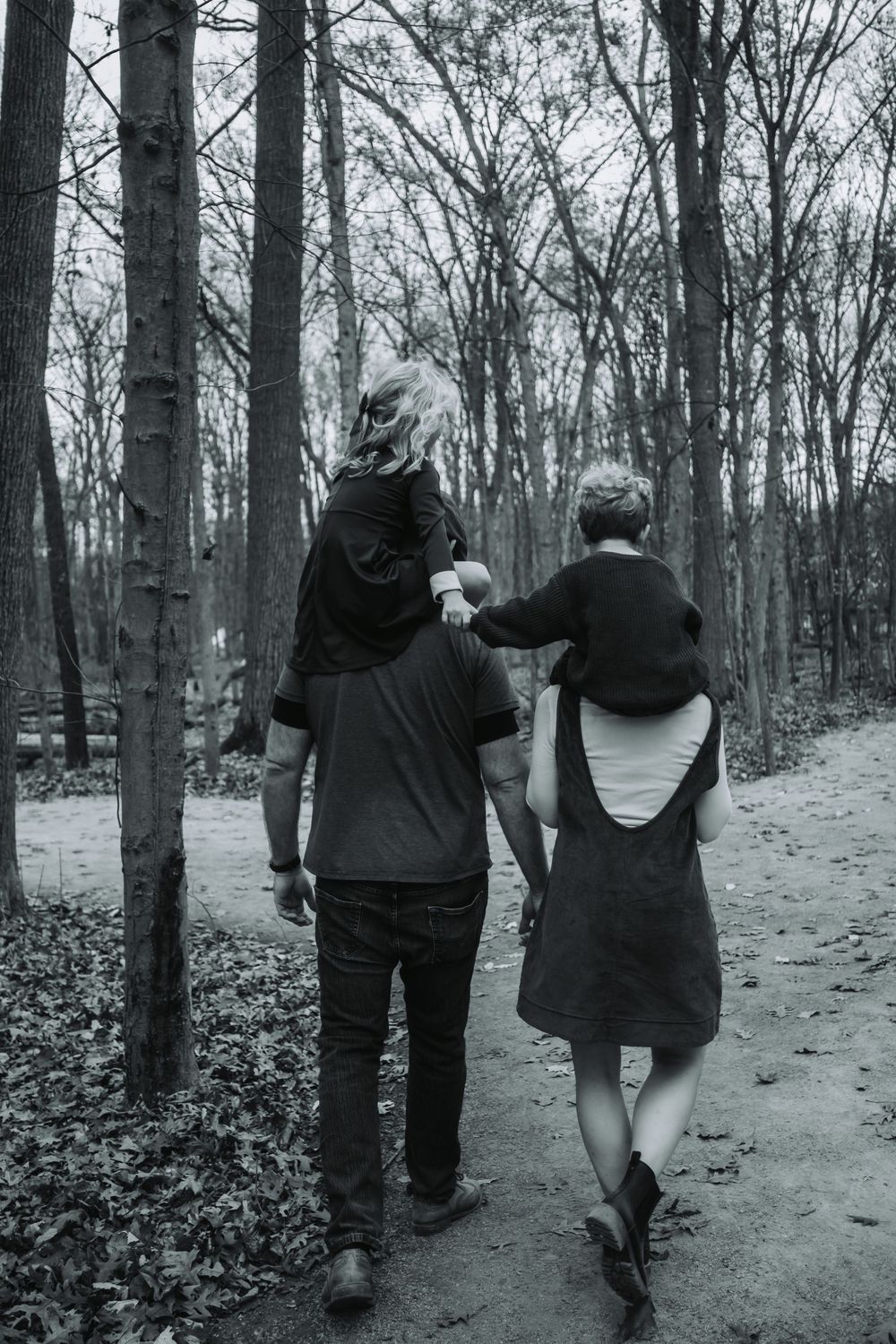 A family of four walk through the woods in black and white. The kids are holding hand while riding on their parents’ shoulders.