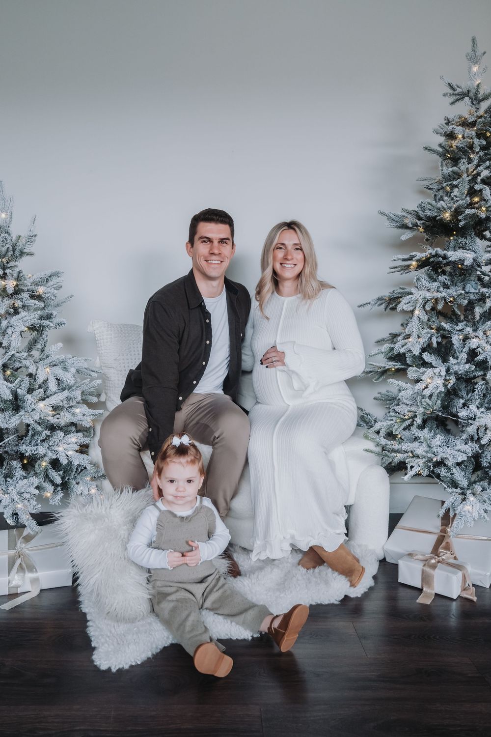 A family of 3 sits on a bench next to two white Christmas trees. The mother is pregnant.