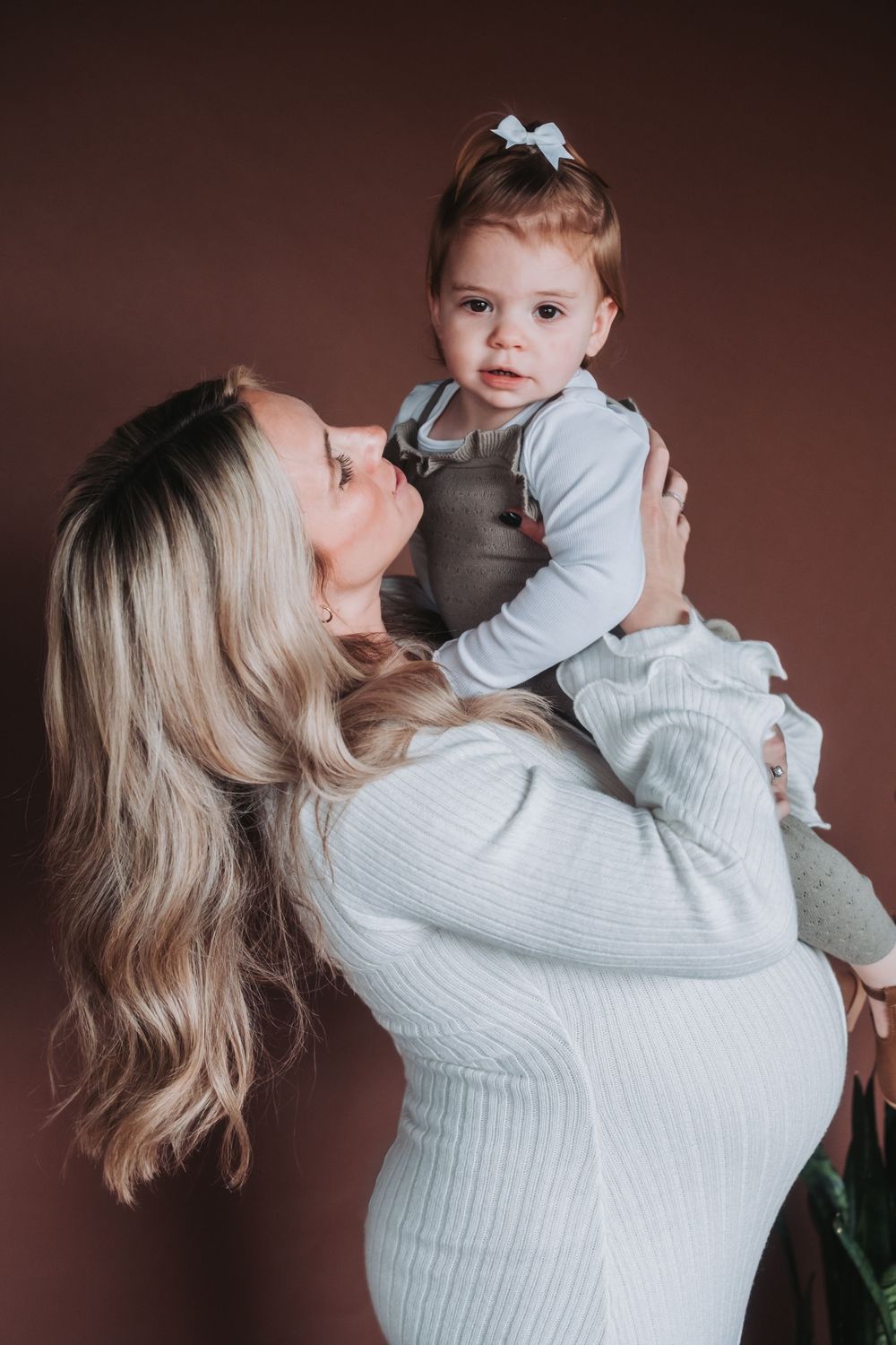A pregnant mother holds her daughter and admires her face, which is turned toward the camera.
