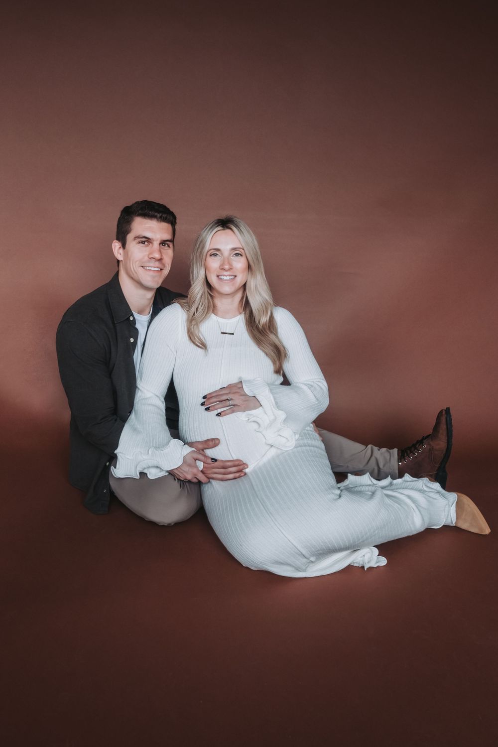 A couple sits together on a brown backdrop as the husband holds his wife’s pregnant belly.