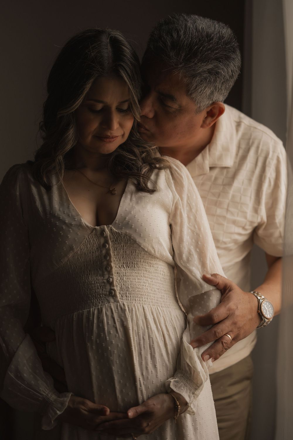 Intimate maternity photo of couple in white clothing embracing in soft natural window light.