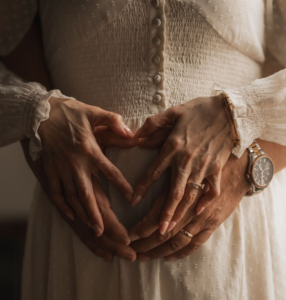 A person's hands forming a heart shape on their stomach while wearing a white lace dress and vintage watch.