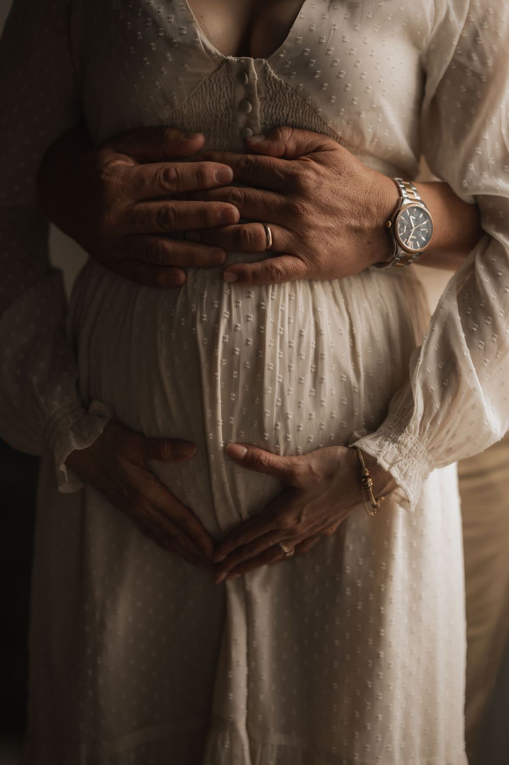 Close-up of hands tenderly cradling a pregnant belly while wearing a flowing white dress and wristwatch.