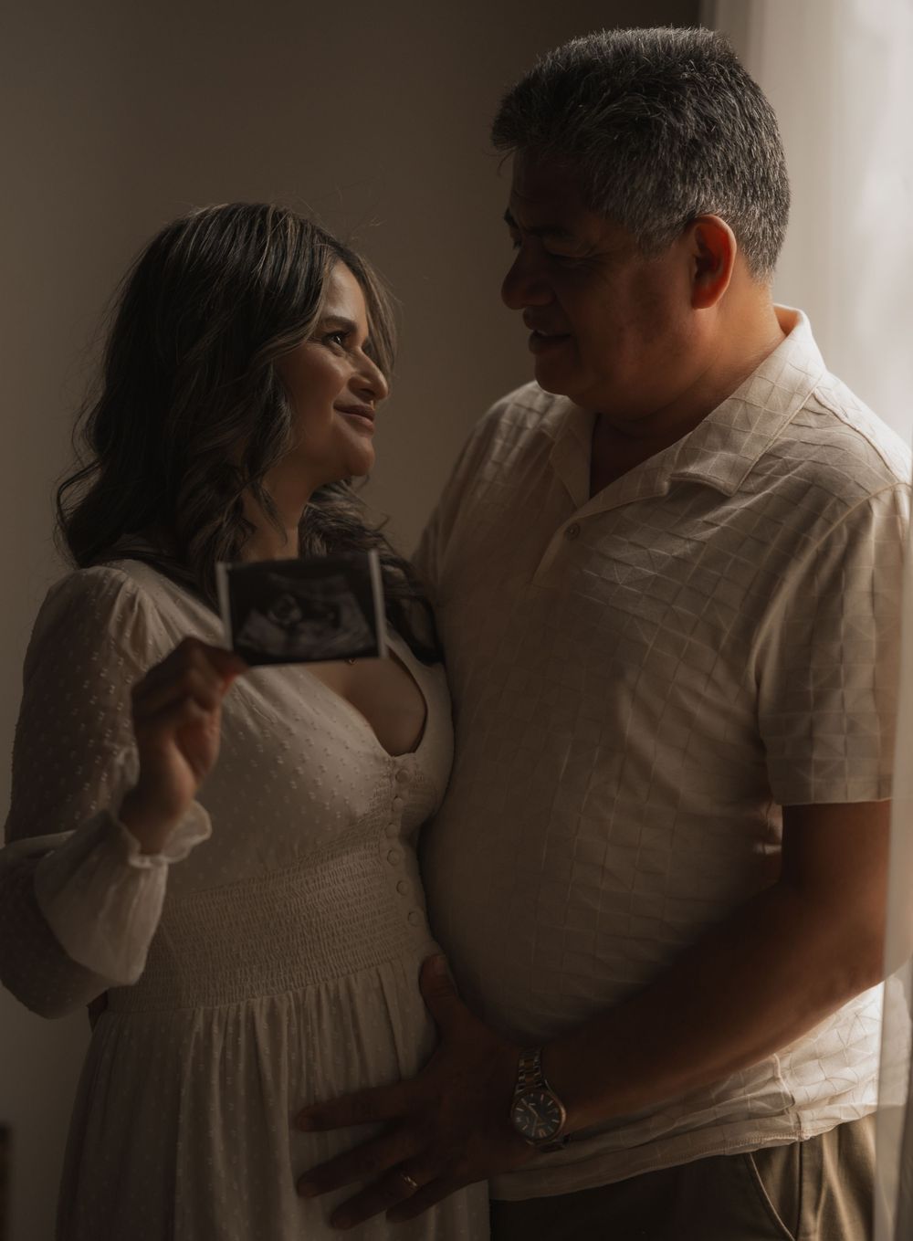 Couple in white clothing shares intimate moment while holding ultrasound photo in dimly lit setting.