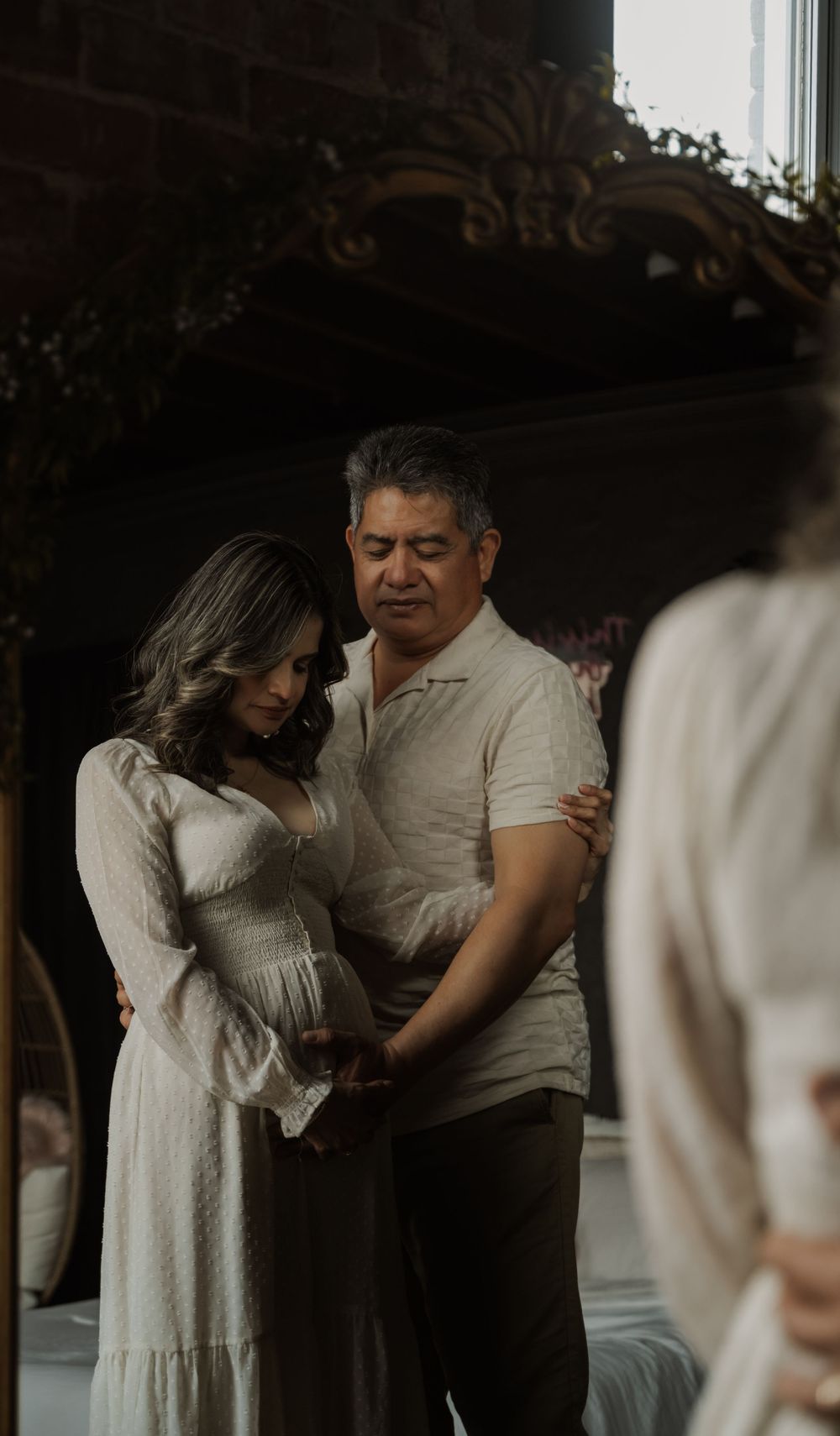 A couple embraces tenderly during a maternity photoshoot, with the pregnant woman wearing a flowing white dress.