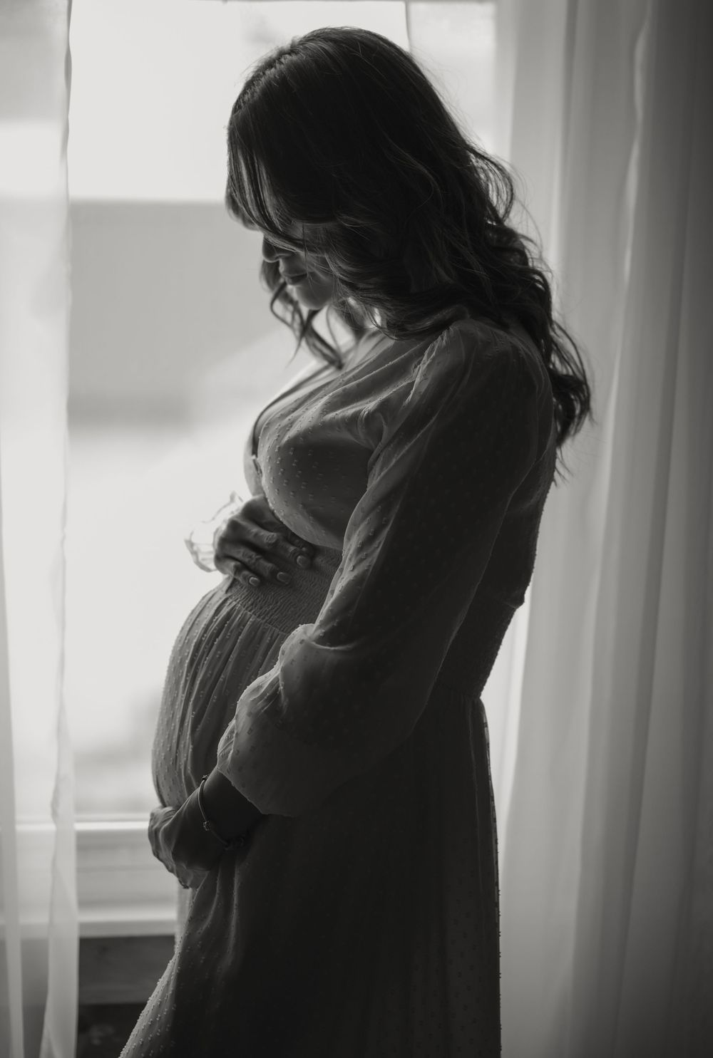 A black and white silhouette of an expectant mother standing by a window in a contemplative pose wearing a flowing dress.