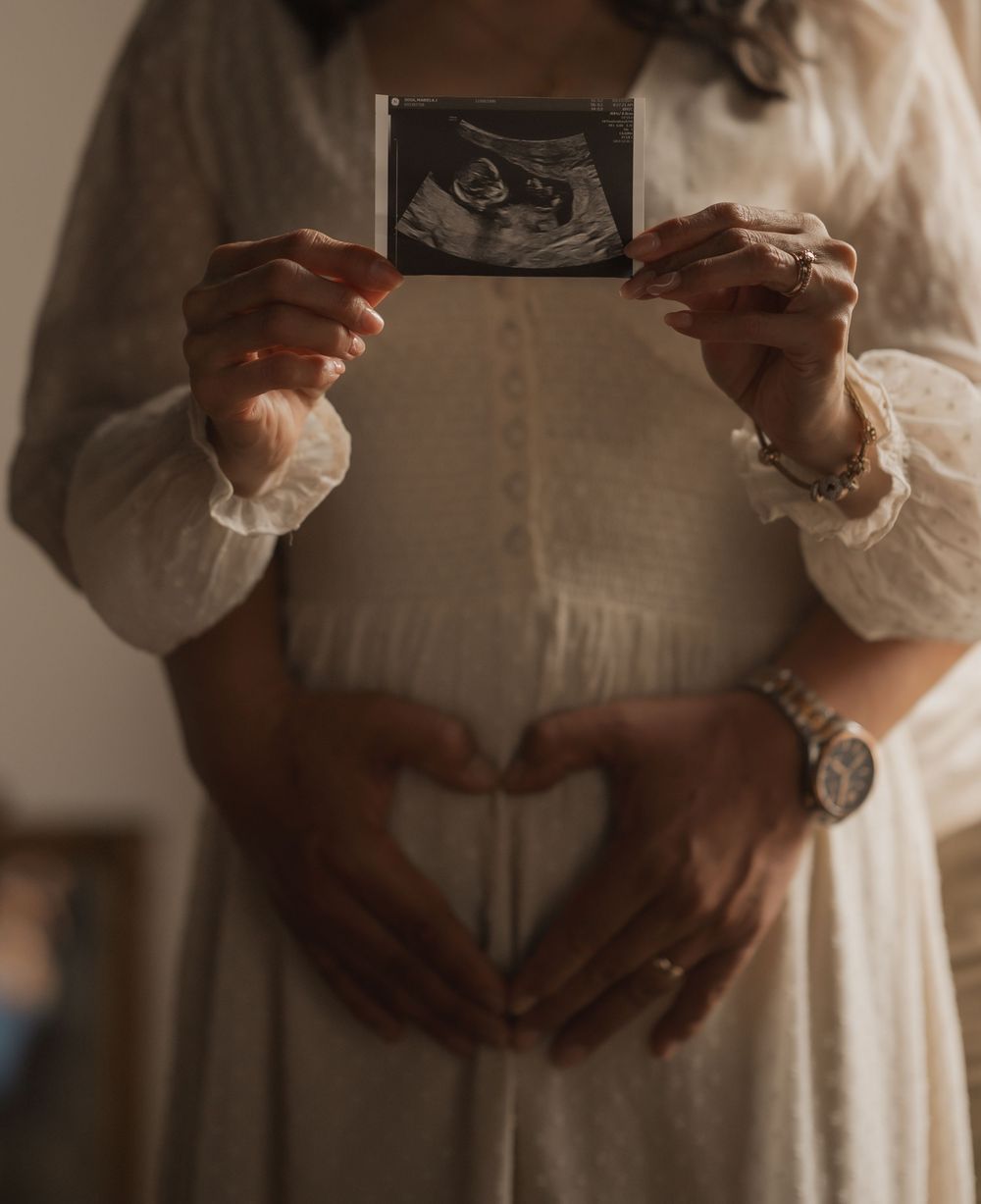 Hands forming heart shape on pregnant belly while holding ultrasound photo in soft natural light.