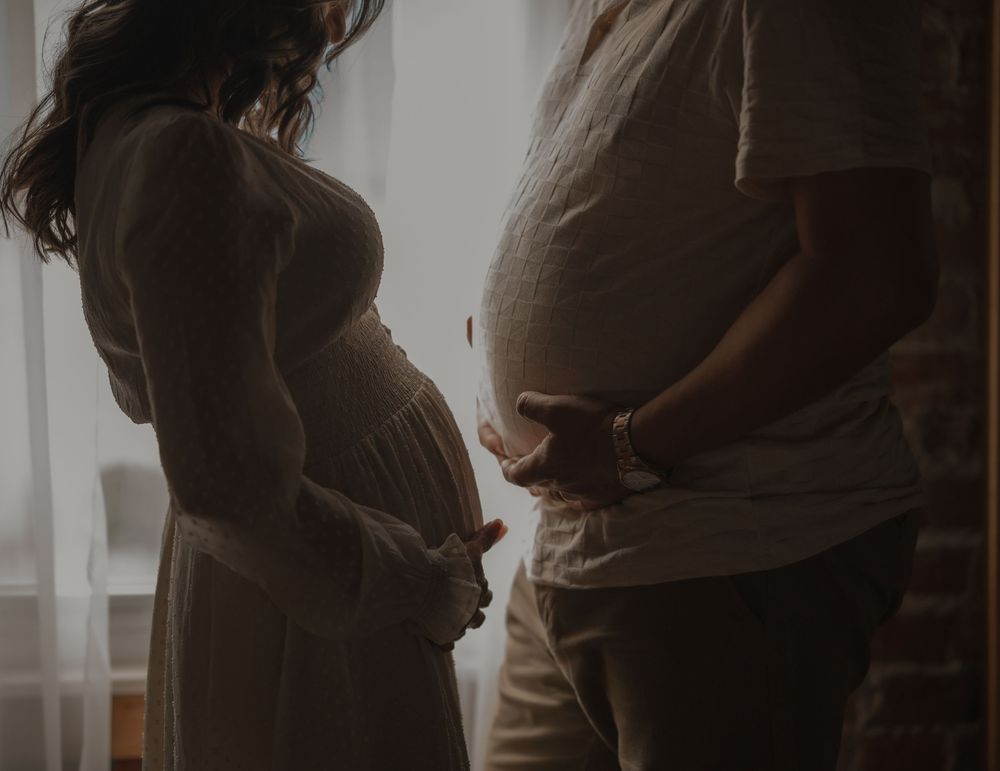 Two pregnant people standing together in silhouette by a window, showing their baby bumps in a tender moment.