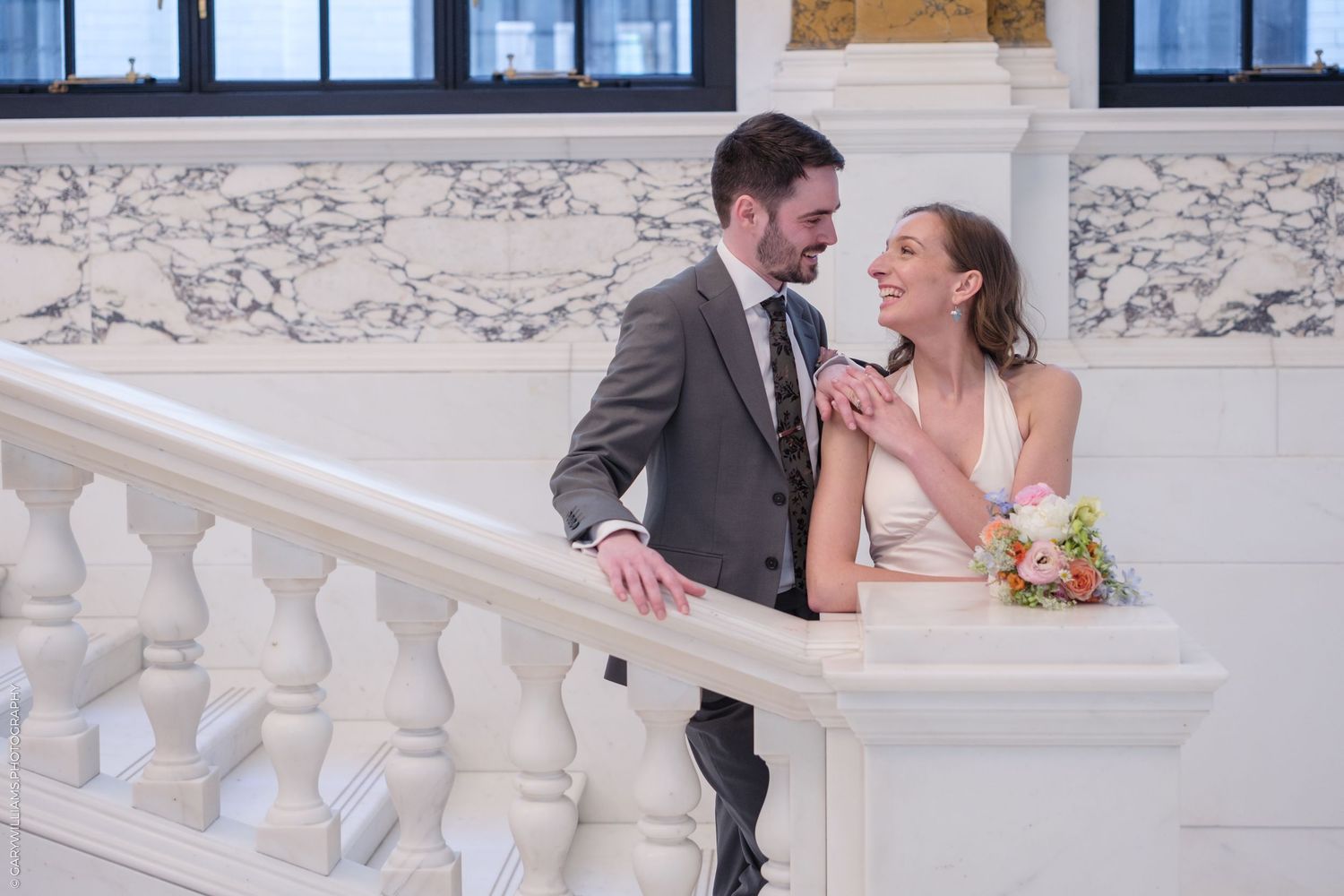 Joyful couple shares a laugh while standing on an elegant white marble staircase with ornate balustrades.