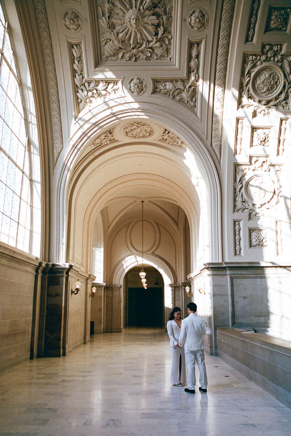 Two people in white stand in a grand classical corridor with ornate ceiling details.
