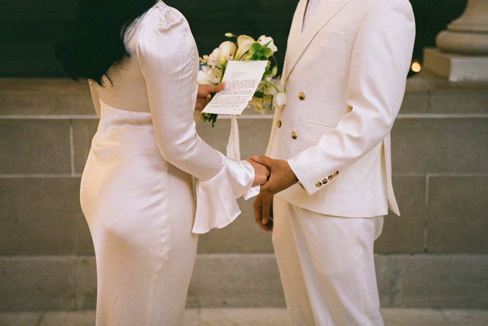 Two people in white formal attire exchange documents during an intimate moment.